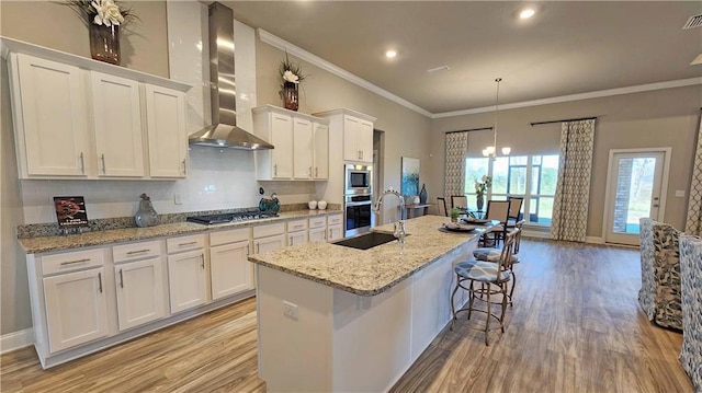 kitchen featuring wall chimney range hood, appliances with stainless steel finishes, a center island with sink, white cabinets, and ornamental molding