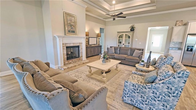 living room with a raised ceiling, crown molding, a brick fireplace, ceiling fan, and light wood-type flooring