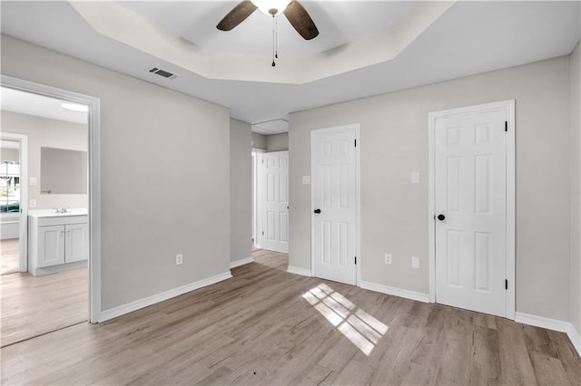 unfurnished bedroom featuring a raised ceiling, ensuite bathroom, light hardwood / wood-style flooring, and ceiling fan