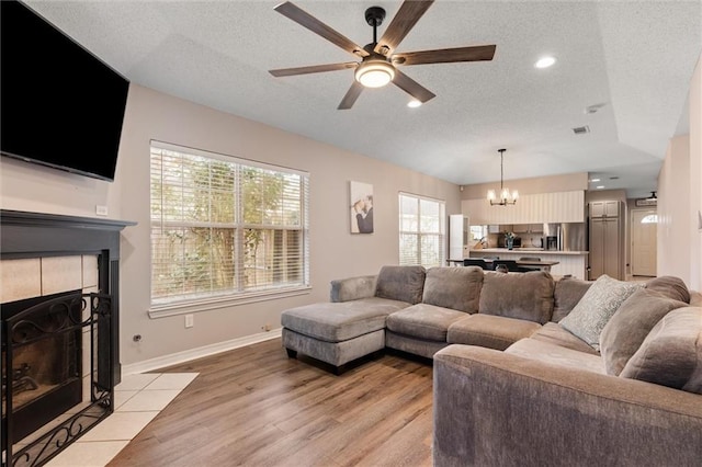 living area with baseboards, light wood finished floors, a textured ceiling, a tiled fireplace, and ceiling fan with notable chandelier