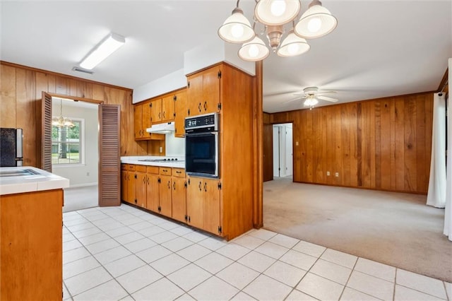 kitchen featuring ceiling fan with notable chandelier, wooden walls, wall oven, light carpet, and black refrigerator