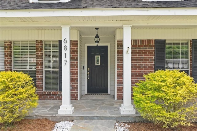 doorway to property with brick siding, a porch, and a shingled roof