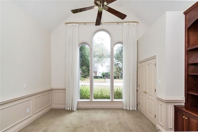 foyer with ceiling fan, light colored carpet, vaulted ceiling, wainscoting, and a decorative wall