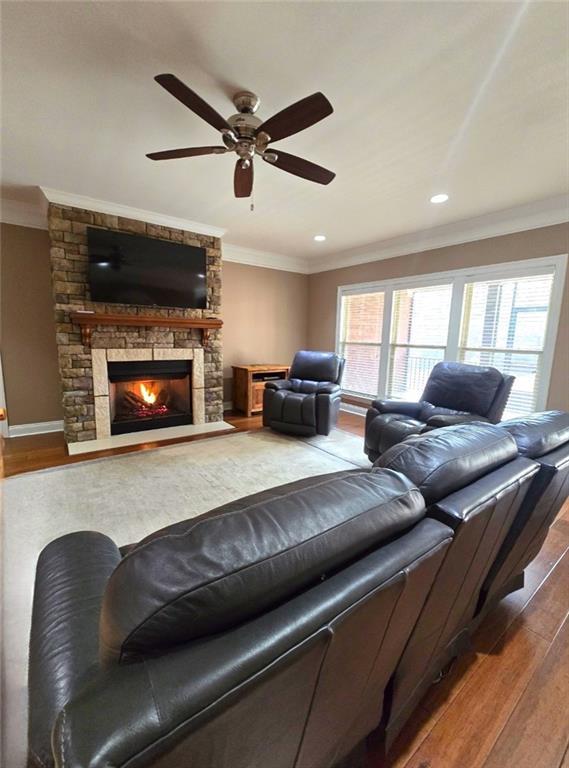 living room with ceiling fan, a stone fireplace, crown molding, and wood-type flooring