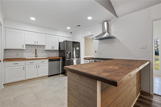 kitchen with butcher block counters, appliances with stainless steel finishes, wall chimney range hood, white cabinetry, and a sink