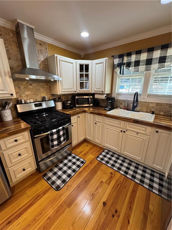 kitchen with butcher block counters, stainless steel appliances, crown molding, wall chimney range hood, and a sink