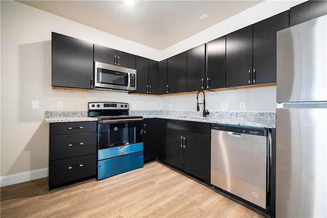 kitchen featuring dark cabinetry, light stone countertops, light wood finished floors, a sink, and appliances with stainless steel finishes