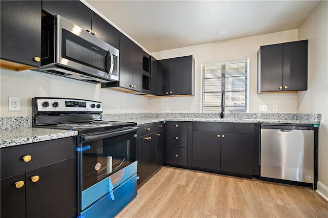 kitchen featuring appliances with stainless steel finishes, light wood-type flooring, dark cabinetry, and light stone countertops