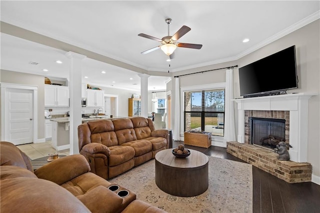 living room with decorative columns, light wood-style flooring, ceiling fan, ornamental molding, and a brick fireplace