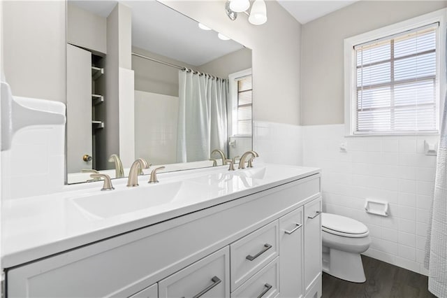 bathroom with double vanity, toilet, a wainscoted wall, wood finished floors, and tile walls