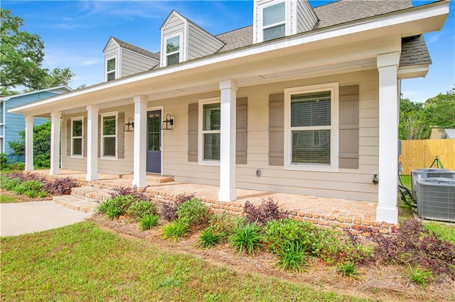 new england style home with central air condition unit, fence, covered porch, and a shingled roof