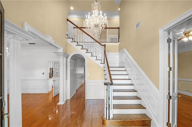 staircase with a towering ceiling, ornate columns, crown molding, wood-type flooring, and a notable chandelier