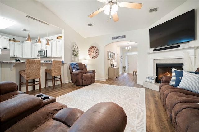 living room with ceiling fan with notable chandelier, wood-type flooring, a tile fireplace, and vaulted ceiling