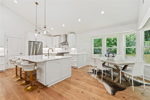 kitchen with stainless steel refrigerator, wall chimney exhaust hood, light hardwood / wood-style flooring, a kitchen island with sink, and white cabinets