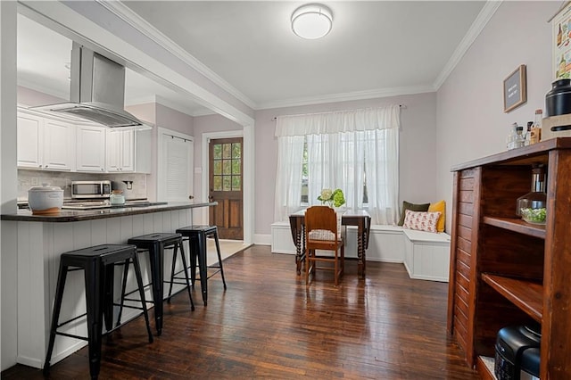 kitchen featuring dark countertops, a breakfast bar area, stainless steel microwave, wall chimney exhaust hood, and dark wood-style floors