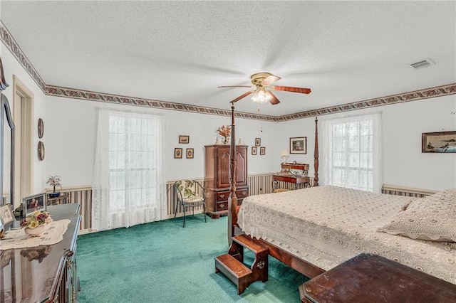 carpeted bedroom with visible vents, a wainscoted wall, ceiling fan, and a textured ceiling