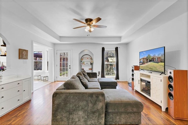 living room featuring a ceiling fan, a tray ceiling, arched walkways, and wood finished floors