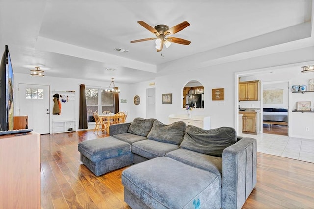 living room with arched walkways, a raised ceiling, visible vents, light wood-style flooring, and ceiling fan with notable chandelier