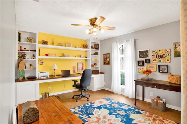 office area featuring light wood finished floors, baseboards, a ceiling fan, a textured ceiling, and built in desk