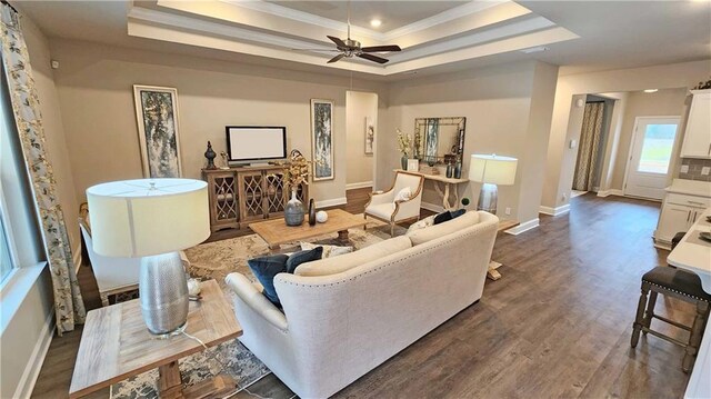living area featuring ceiling fan, baseboards, dark wood-type flooring, a tray ceiling, and ornamental molding