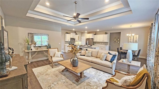 living room featuring ornamental molding, ceiling fan with notable chandelier, baseboards, a tray ceiling, and dark wood-style floors