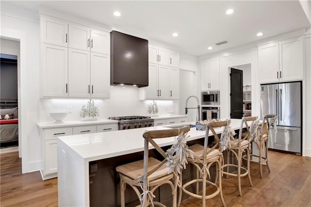 kitchen with wall chimney exhaust hood, a breakfast bar, stainless steel appliances, a center island with sink, and light hardwood / wood-style floors