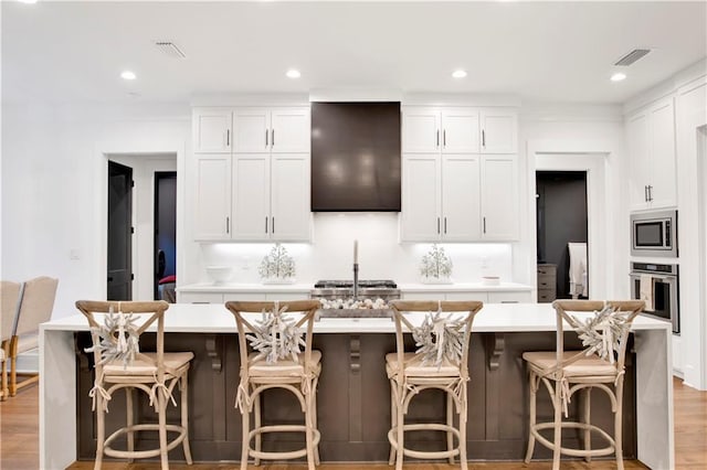 kitchen featuring stainless steel appliances, a kitchen bar, a kitchen island with sink, white cabinets, and exhaust hood