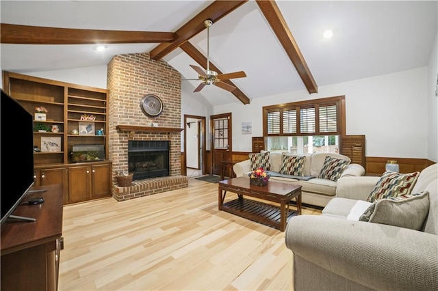 living room with a wainscoted wall, a fireplace, lofted ceiling with beams, light wood-style floors, and a ceiling fan