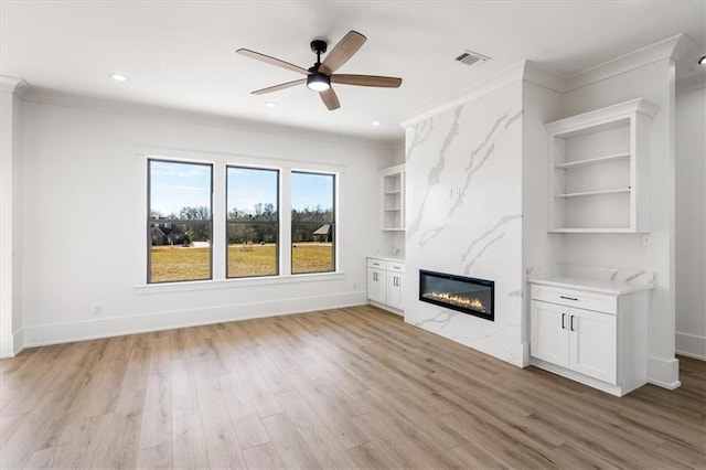 unfurnished living room with a large fireplace, ceiling fan, crown molding, built in features, and light hardwood / wood-style floors