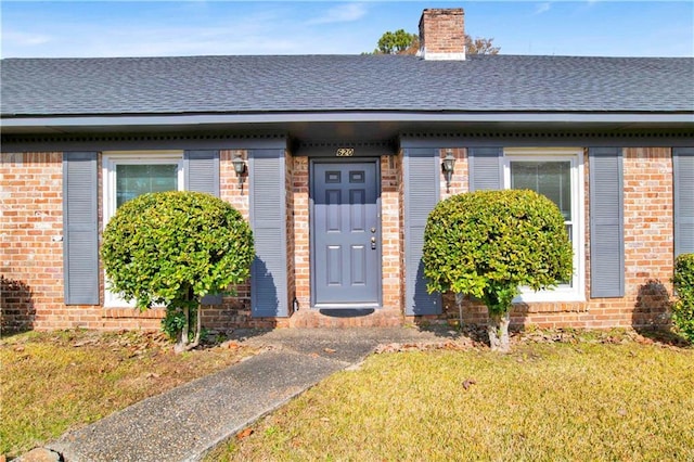 entrance to property with brick siding, roof with shingles, a chimney, and a lawn