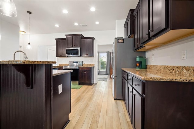 kitchen with stainless steel appliances, dark brown cabinets, light wood-style floors, light stone counters, and a kitchen breakfast bar
