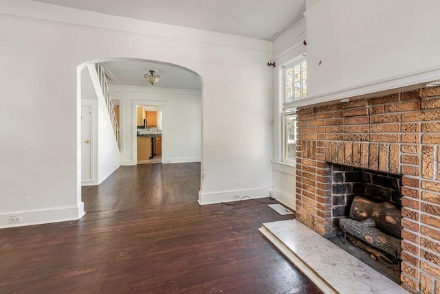 unfurnished living room featuring a brick fireplace, arched walkways, baseboards, and wood finished floors