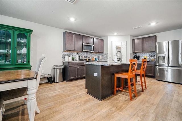 kitchen with a breakfast bar, light hardwood / wood-style flooring, appliances with stainless steel finishes, an island with sink, and dark brown cabinets