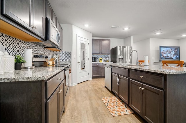 kitchen with appliances with stainless steel finishes, tasteful backsplash, a kitchen island with sink, dark brown cabinets, and light hardwood / wood-style flooring