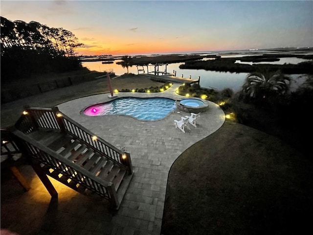 pool at dusk featuring a water view, a patio area, an outdoor pool, and an in ground hot tub