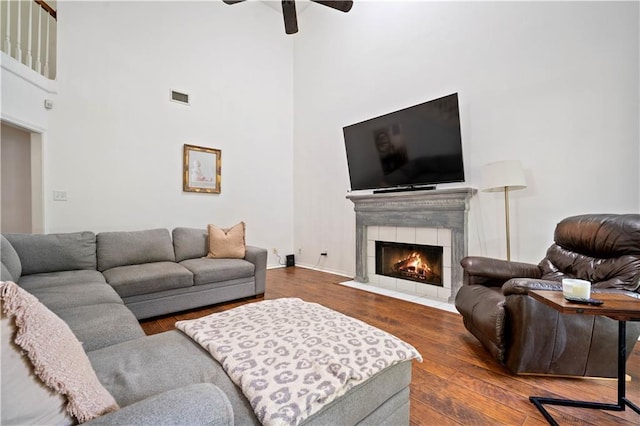 living area featuring a tiled fireplace, wood finished floors, ceiling fan, visible vents, and a high ceiling