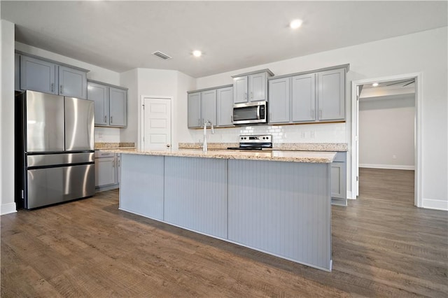 kitchen featuring a kitchen island with sink, gray cabinets, dark wood-type flooring, backsplash, and appliances with stainless steel finishes