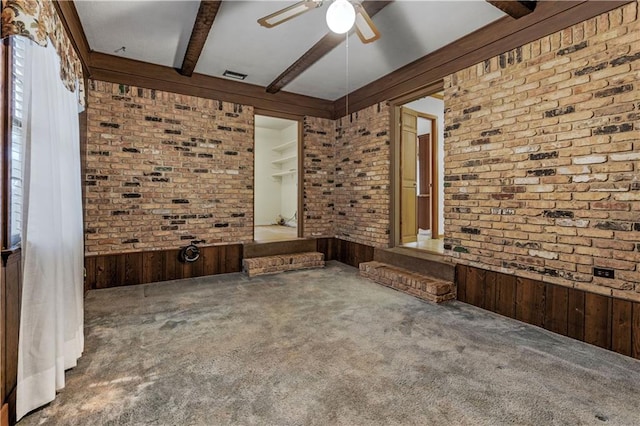 carpeted empty room featuring beamed ceiling, wooden walls, ceiling fan, and brick wall