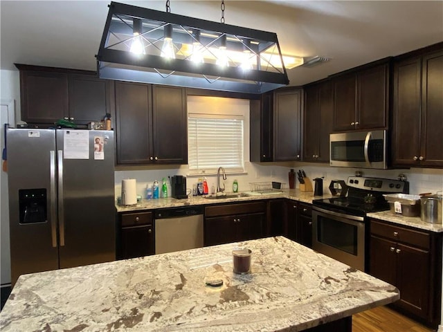 kitchen with a sink, light stone counters, dark brown cabinetry, hanging light fixtures, and appliances with stainless steel finishes