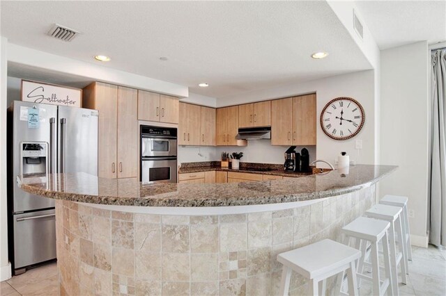 kitchen with appliances with stainless steel finishes, light brown cabinetry, dark stone counters, a peninsula, and recessed lighting