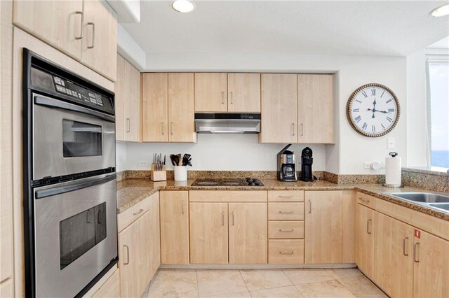 kitchen with light brown cabinetry, under cabinet range hood, double oven, black electric stovetop, and light stone countertops