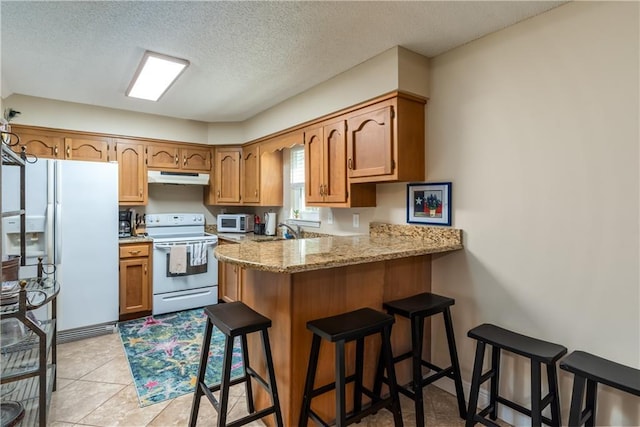 kitchen with under cabinet range hood, a peninsula, white appliances, a kitchen breakfast bar, and brown cabinets