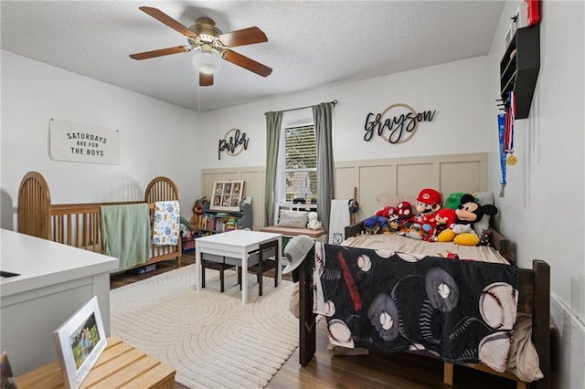 bedroom featuring a textured ceiling, wood finished floors, ceiling fan, wainscoting, and a decorative wall