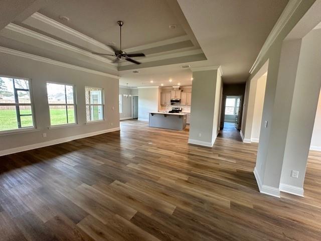 unfurnished living room featuring a raised ceiling, ceiling fan, ornamental molding, and dark hardwood / wood-style flooring