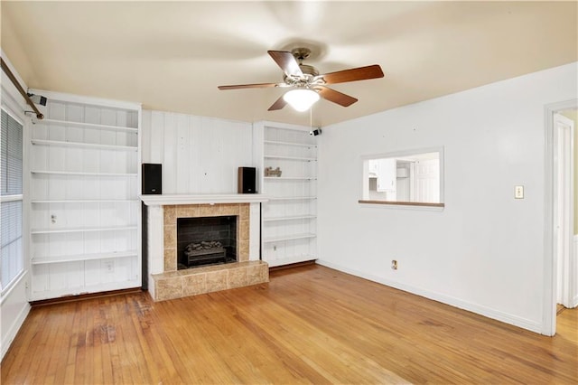 unfurnished living room featuring a tile fireplace, wood-type flooring, ceiling fan, and built in shelves