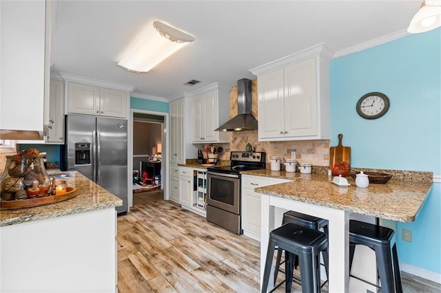 kitchen with light stone countertops, stainless steel appliances, wall chimney range hood, white cabinets, and light hardwood / wood-style floors