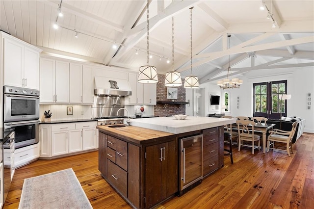 kitchen with dark brown cabinetry, double oven, an island with sink, beverage cooler, and range hood