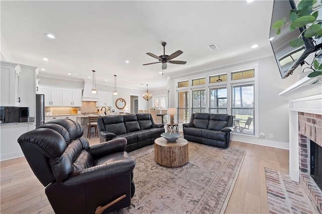 living room featuring light wood-style flooring, ceiling fan with notable chandelier, visible vents, ornamental molding, and a brick fireplace