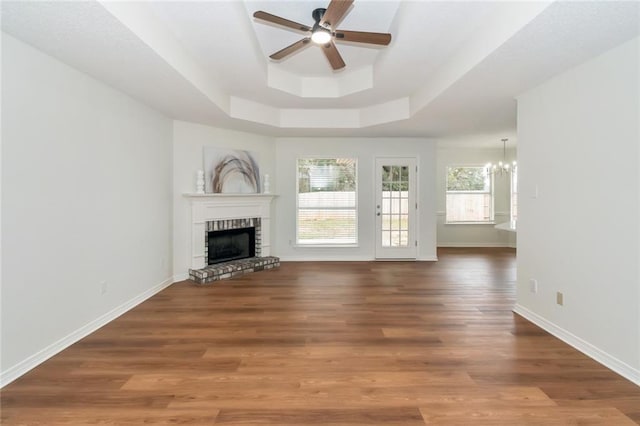 unfurnished living room with a tray ceiling, a brick fireplace, wood finished floors, and ceiling fan with notable chandelier