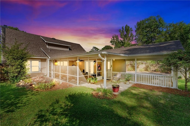 back of house at dusk featuring roof with shingles, a yard, a patio area, and a sunroom